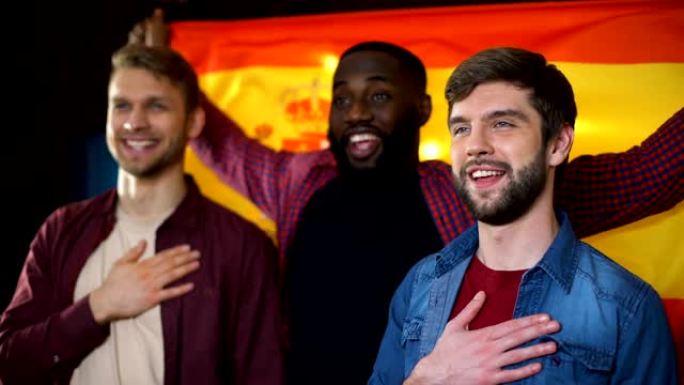 西班牙多民族男性足球迷唱着国歌，挥舞着旗帜