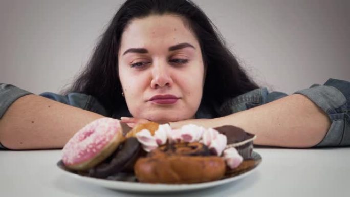 胖胖的黑发白人女孩坐在盘子前拿着糖果的特写脸。饥饿丰满的女人看着美味的甜点，舔嘴唇。超重，医疗保健，