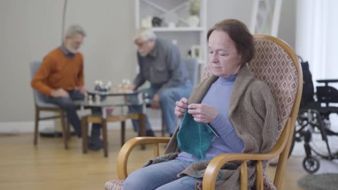 白人老妇人在扶手椅上编织，模糊的男人在背景下下棋。住在养老院的老年退休人员。