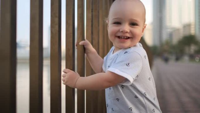 1岁那年的笑男孩，抱着栏杆跳舞，学习走路，迈出第一步，穿着白色连身裤站在海滨