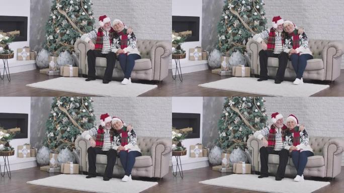 快乐的高加索老年夫妇的肖像坐在圣诞树旁边的沙发上，看着相机，微笑着。丈夫抱着可爱的妻子说话。电影院4