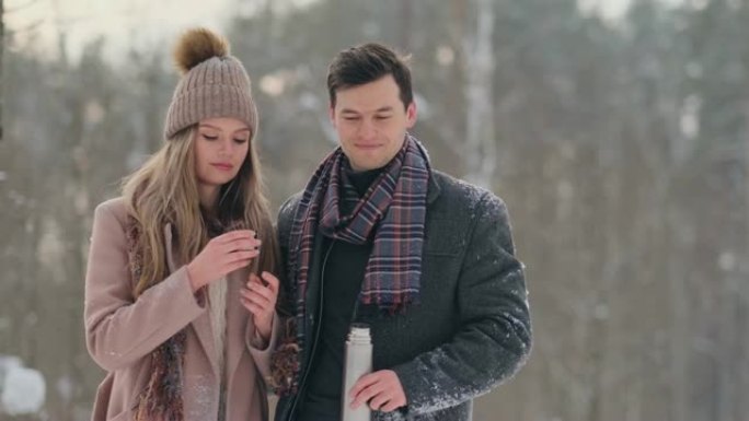 冬天森林里一对相爱的男女在热水瓶里喝茶。冬天在公园里穿着外套的时尚男人和女人散步。慢动作
