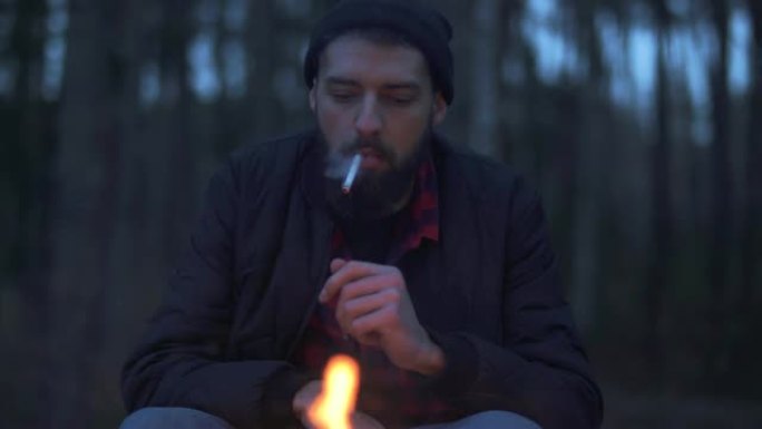 坐在森林火炉旁的大胡子家伙开始抽烟。野蛮的未刮胡子的家伙在森林里的火旁休息。