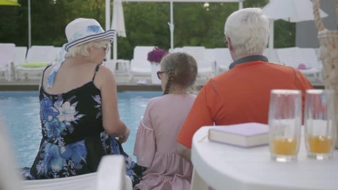 祖母，祖父和孙子匡威坐在豪华游泳池的边缘。
