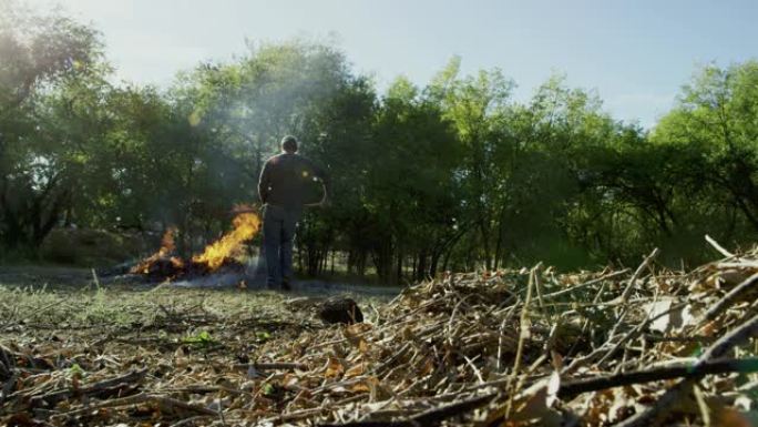 一个四十多岁的高加索人用干草叉捡起木棍和树枝，然后将它们扔到户外的一个大火烧堆上