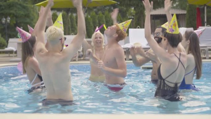 阳光明媚的户外，戴着派对帽的快乐年轻人在泳池里围着红发人跳舞。一群快乐的朋友在度假村庆祝生日。游客开