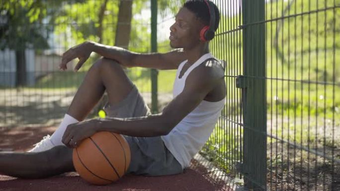 积极的非洲裔美国运动员坐在篮球场上带球，戴着耳机听音乐的侧视图。英俊的年轻人喜欢在阳光明媚的夏日休息