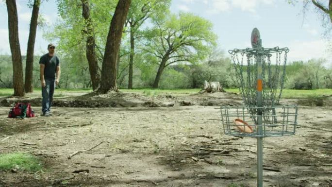 一个四十多岁的高加索人使用反手推杆将圆盘高尔夫球推杆扔进圆盘高尔夫球篮，然后在阳光明媚的日子在没有树