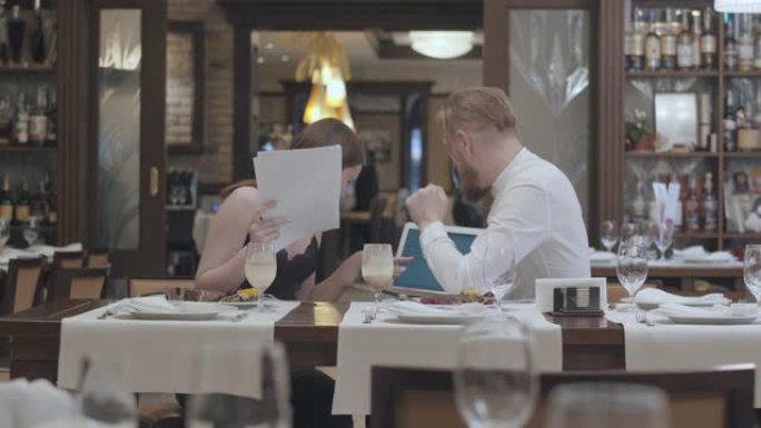 肖像可爱的年轻女子坐在餐厅的桌子上，和一个自信的大胡子男人坐在桌子上。