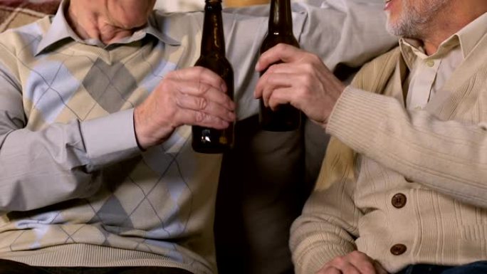 两个养老金领取者碰杯喝啤酒坐在沙发上，空闲时间