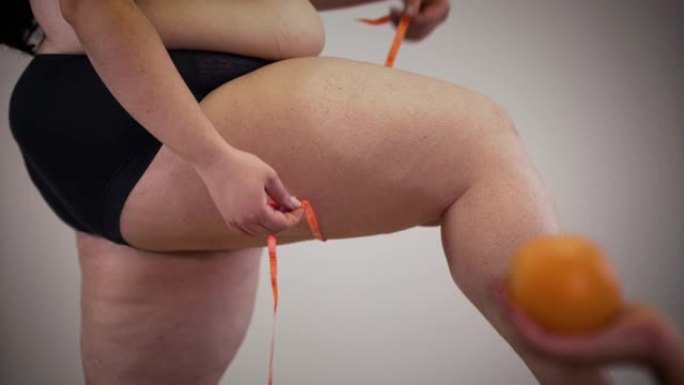 肥胖的高加索人无法辨认的女人用卷尺测量腿，因为女性的手在前景处拿着橙色。专注于背景。超重，肥胖，不健