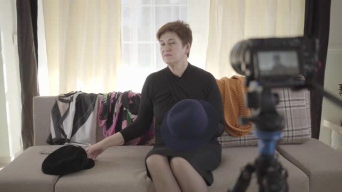 正面的白人女性肖像在相机帽子和围巾上展示。成熟的女人坐在沙发上，为互联网页面录制视频。社交媒体，技术