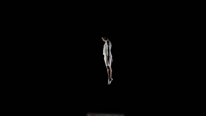 穿着白色衣服的黑色背景的男性体操运动员从下方飞入框架，在空中旋转并以慢动作翻转。比赛中的体操运动员。