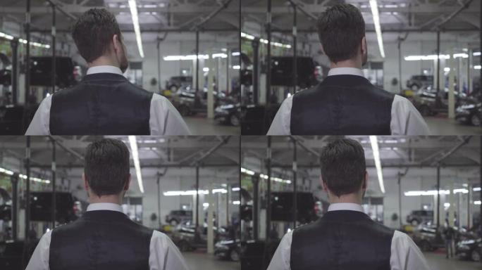 穿着背心和衬衫的高加索人站在修理厂看着汽车的后视图。自信的年轻商人在等他的车修好。保修、服务、汽车维