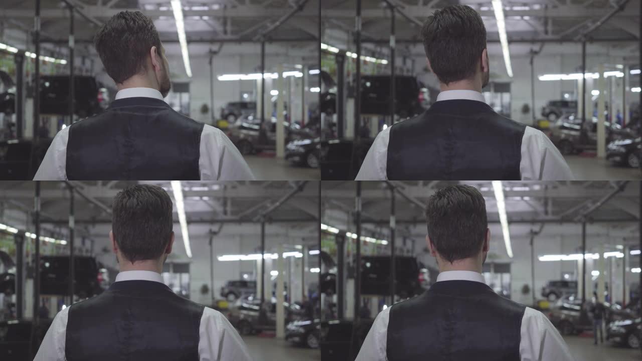 穿着背心和衬衫的高加索人站在修理厂看着汽车的后视图。自信的年轻商人在等他的车修好。保修、服务、汽车维
