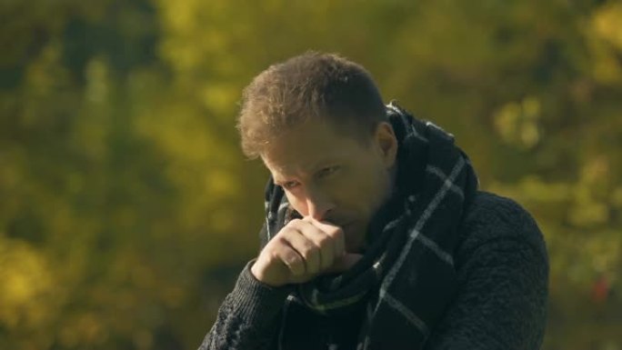生病的男性在围巾上咳嗽，在秋天公园打喷嚏，感冒，感染