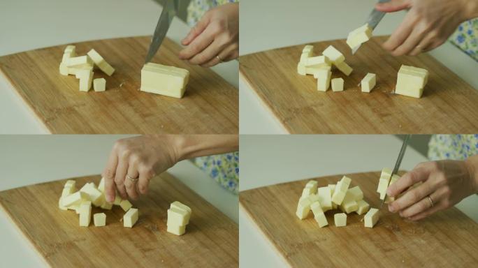 在舒适的室内厨房里，一名年轻女子的手用菜刀在白色桌子上的木制砧板上用菜刀将一根黄油切成小块