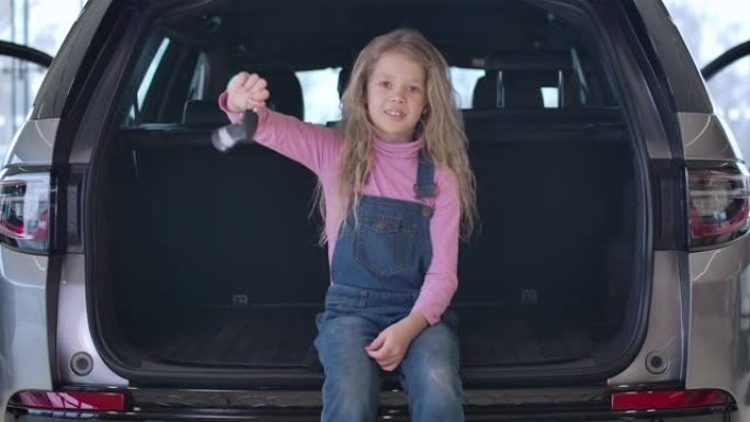 快乐的高加索小女孩坐在汽车后备箱和吹牛钥匙的肖像。迷人的金发碧眼的长卷发孩子在汽车经销店摆姿势。电影