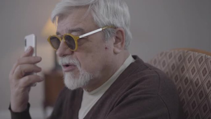 盲人高个子白人戴眼镜在电话里的特写侧视图。在室内使用智能手机的老年男性退休人员。现代科技、生活方式、
