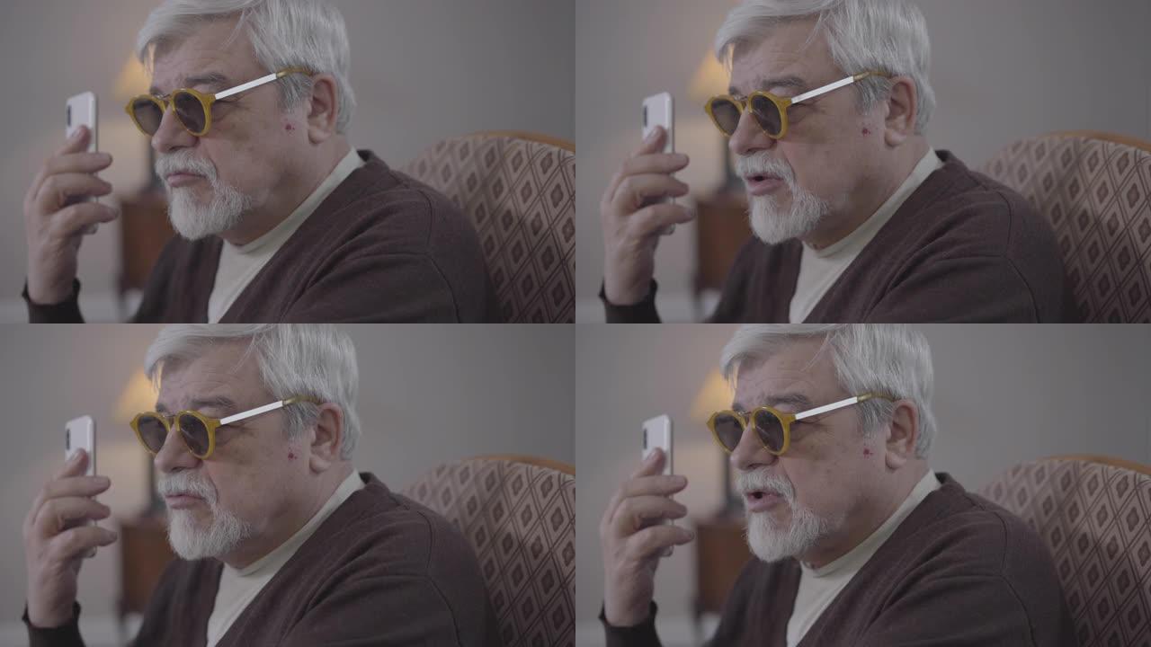 盲人高个子白人戴眼镜在电话里的特写侧视图。在室内使用智能手机的老年男性退休人员。现代科技、生活方式、