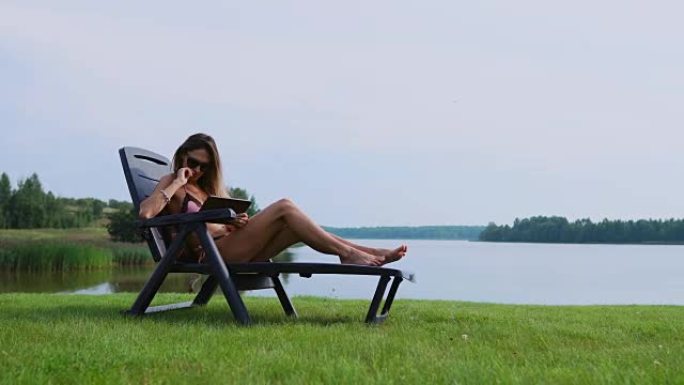 在平板电脑上工作的女人在度假时环游世界，在湖边的海滩上晒日光浴