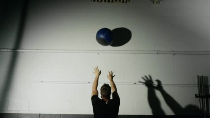 一位二十多岁的有胡子的白人男子表演捡起一个药球，并在灯光明亮的体育馆里进行侧向药球，然后让球掉到地板