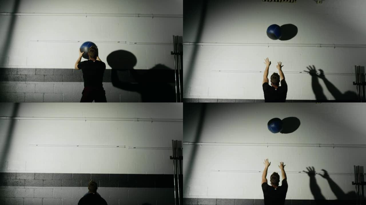 一位二十多岁的有胡子的白人男子表演捡起一个药球，并在灯光明亮的体育馆里进行侧向药球，然后让球掉到地板