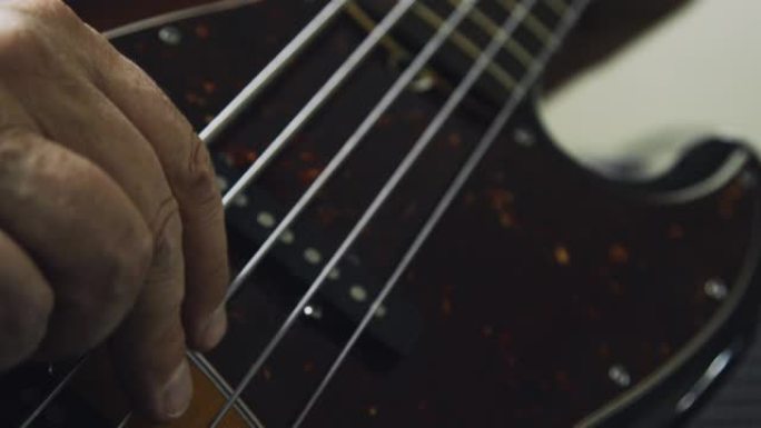 一个五十多岁的高加索人在五弦电贝司吉他的指板上往下压，在室内演奏音乐时拨弦
