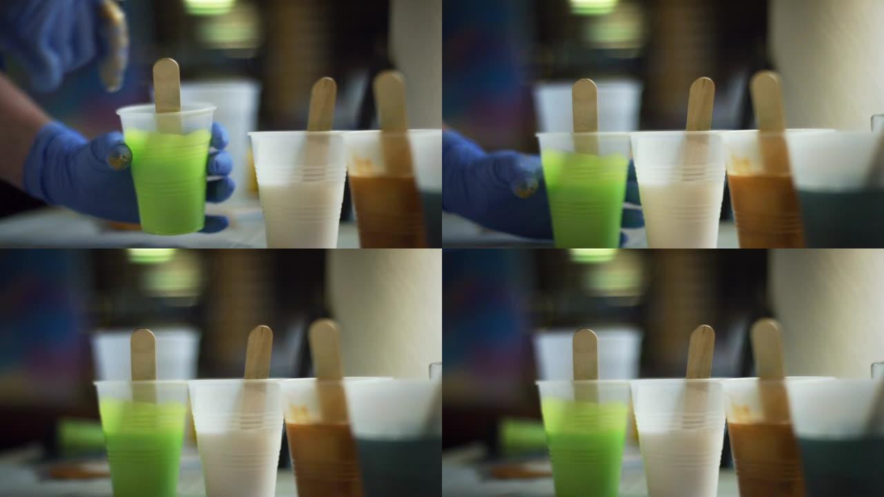 带有安全手套的树脂用木制工艺棒搅动一个塑料杯绿色环氧树脂/树脂，并将其放在艺术工作室中其他几杯有色环