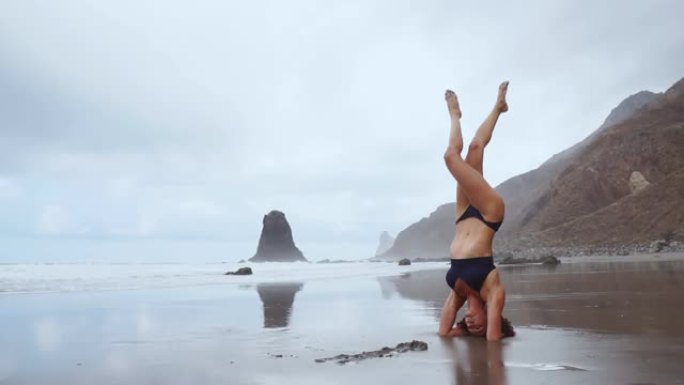 沿着海洋，一个瑜伽倒立冥想的女人。健康的生活方式。站在水边的沙子里平衡你的头
