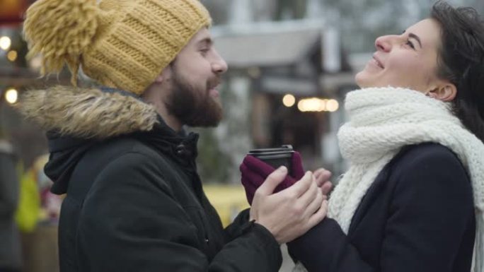欢快的高加索男人和女人站在冬季集市上的咖啡杯旁大笑的侧视图。幸福的年轻家庭一起在户外休息。休闲，生活