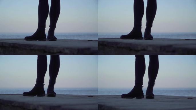 女性的腿站在户外的木制平台上。穿着紧身紧身裤和靴子的女人的腿在水面的背景下。