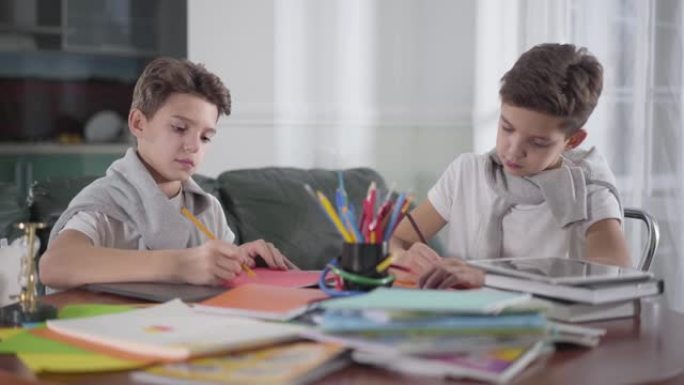 两个聪明的高加索双胞胎兄弟在家里用彩色铅笔画画。男生一起做作业。有创意的兄弟姐妹坐在桌边画画。