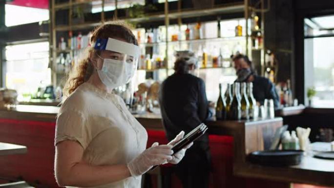 新型冠状病毒肺炎大流行期间穿着PPE的女服务员使用平板电脑看着相机