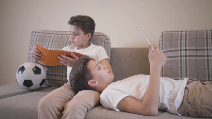 两个白人男生在家躺在沙发上休息。穿着类似衣服的双胞胎兄弟在室内读书。一个男孩使用平板电脑，他的兄弟手