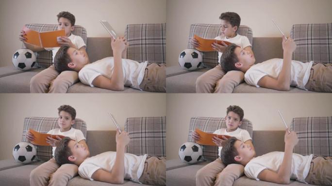 两个白人男生在家躺在沙发上休息。穿着类似衣服的双胞胎兄弟在室内读书。一个男孩使用平板电脑，他的兄弟手