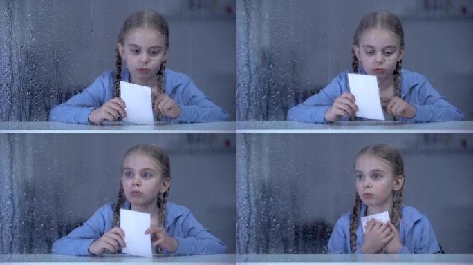 小女孩在下雨的窗户后面看着照片，孤儿失踪的父母