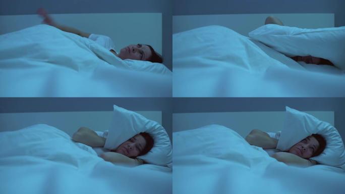 恼怒的女人在床上失眠，用吵闹的邻居的枕头遮住耳朵