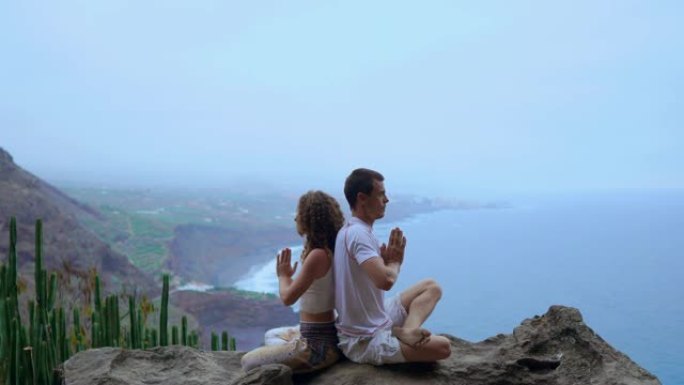男人和女人背靠背坐在山顶上的岩石上冥想，并在海洋的背景下做瑜伽。