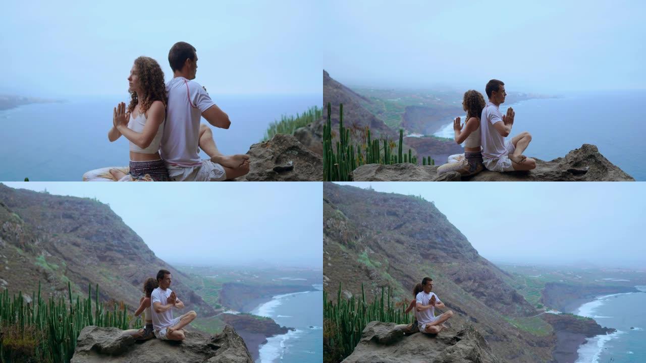男人和女人背靠背坐在山顶上的岩石上冥想，并在海洋的背景下做瑜伽。