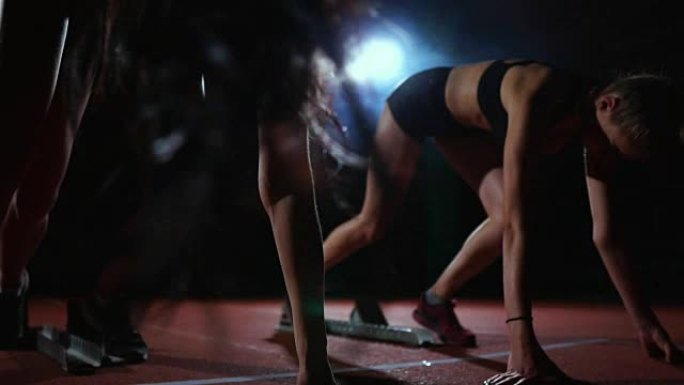 三个穿着黑色衣服的女孩在起跑板上，在灯光下开始比赛，并朝着终点奔跑