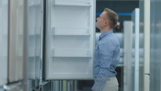 一个穿着蓝色衬衫的年轻人打开电冰箱的门在电器商店，并比较其他型号的新房子