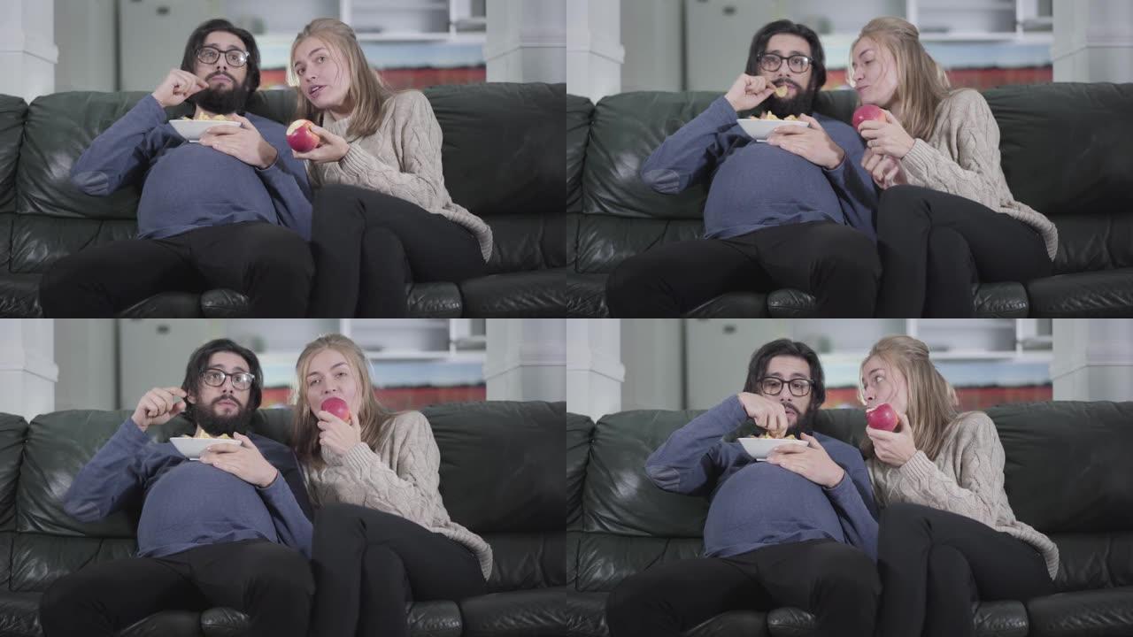 有趣的高加索夫妇坐在沙发上看电视。怀孕的丈夫吃薯片，年轻漂亮的妻子咬苹果。家庭交换社会角色。开玩笑，