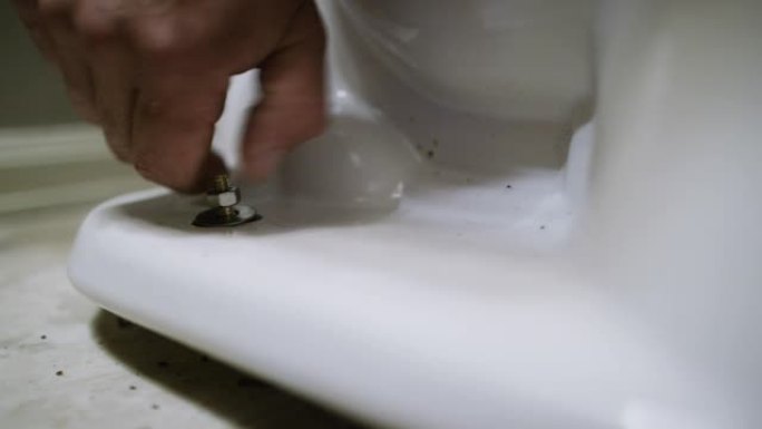 一名高加索修理工将螺母放在法兰螺栓上，然后用手指将其拧紧，以将瓷质马桶固定在室内家用浴室的地板上