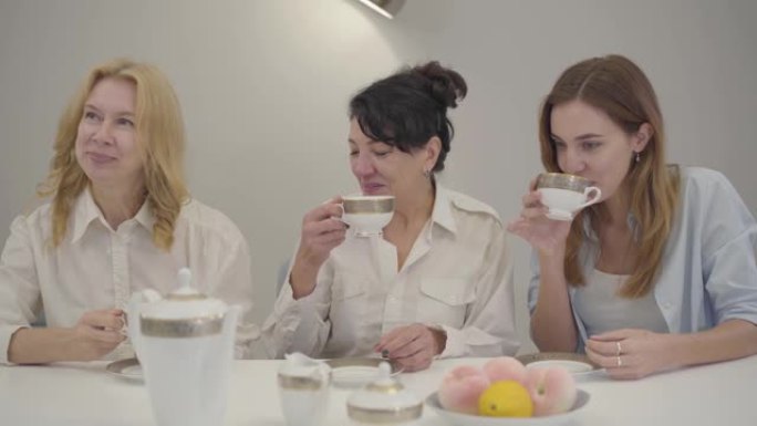 三个不同年龄的白人妇女坐在桌旁喝茶或咖啡。朋友或家人聚集在一起，交谈和微笑。快乐的人在室内休息。