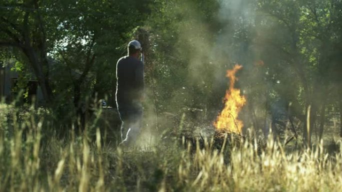 一个四十多岁的高加索人用干草叉捡起木棍和树枝，然后将它们扔到户外的一个大火烧堆上