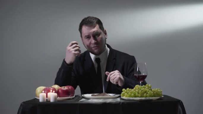 穿着西装的严肃绅士坐在小桌子前的肉，葡萄，苹果，红酒和蜡烛的宁静。