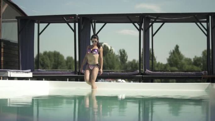 穿着闪亮银色泳衣的黑发女人穿着运动的身体进入游泳池的水中。穿着比基尼的孤独女士的休闲。