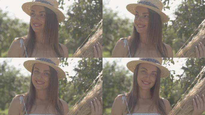 戴着草帽和白色长裙的可爱年轻女子站在绿色的夏日花园里，手里拿着一束干草。