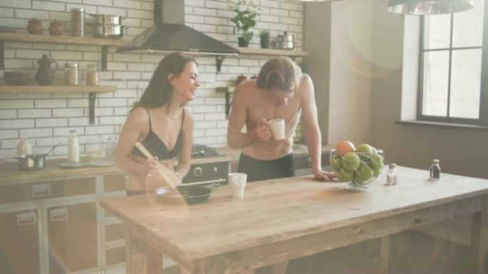 一对年轻夫妇在漂亮的阁楼厨房里微笑着。一个男孩和一个女孩穿着内衣，站在厨房桌子旁边的阳光下喝着他们的